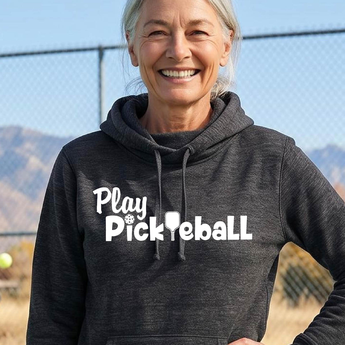A smiling woman outdoors wearing a dark charcoal heather cowl-neck pullover hoodie with the white text 'Play Pickleball' across the chest, photographed in front of a fence and hills.
