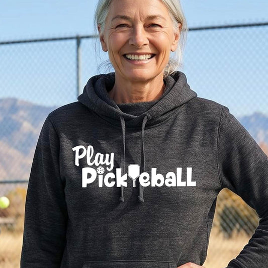 A smiling woman outdoors wearing a dark charcoal heather cowl-neck pullover hoodie with the white text 'Play Pickleball' across the chest, photographed in front of a fence and hills.
