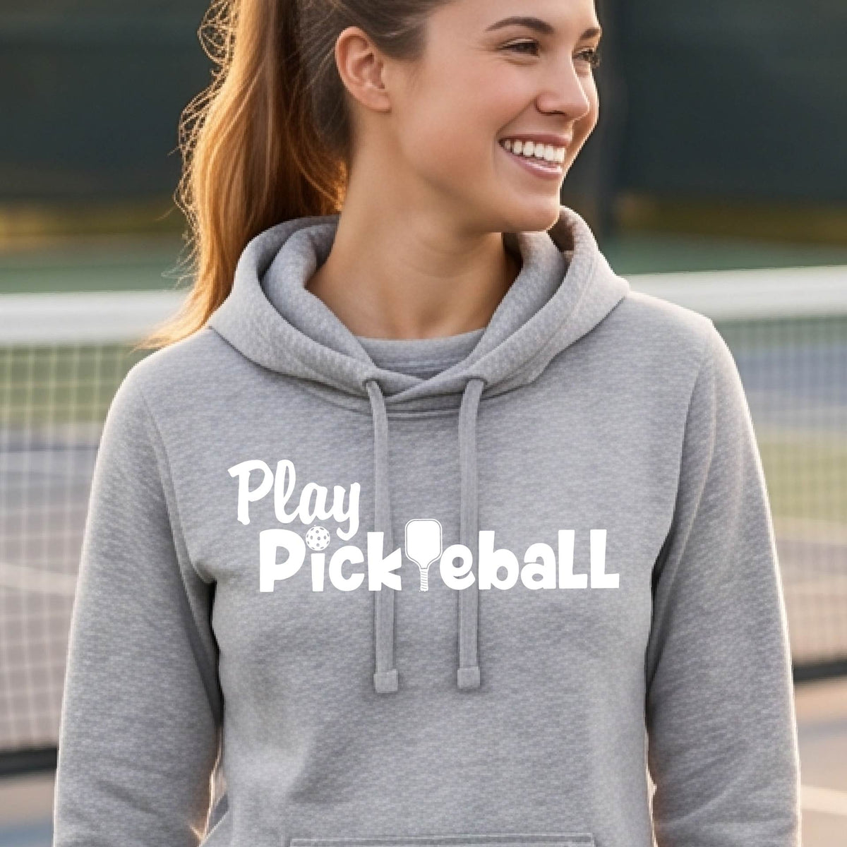 A woman wearing a heather gray hoodie with 'Play Pickleball' printed in white across the chest, standing on an outdoor pickleball court.