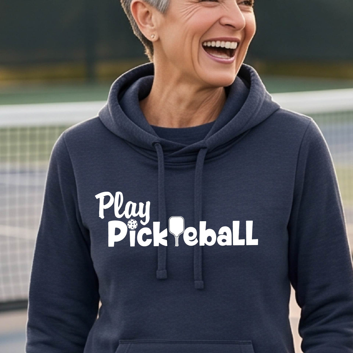 A smiling woman outdoors wearing a dark charcoal heather cowl-neck pullover hoodie with the white text 'Play Pickleball' across the chest, photographed in front of a fence and hills.