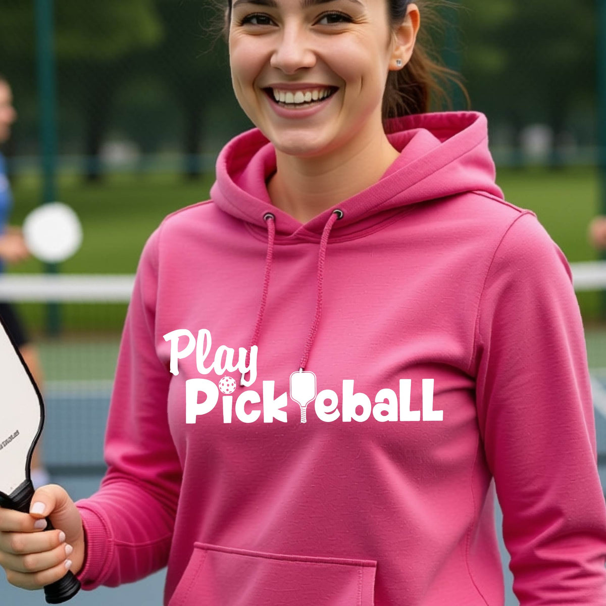 A smiling woman wearing a bright pink hoodie with the text 'Play Pickleball' printed in white across the chest, holding a pickleball paddle on an outdoor court.