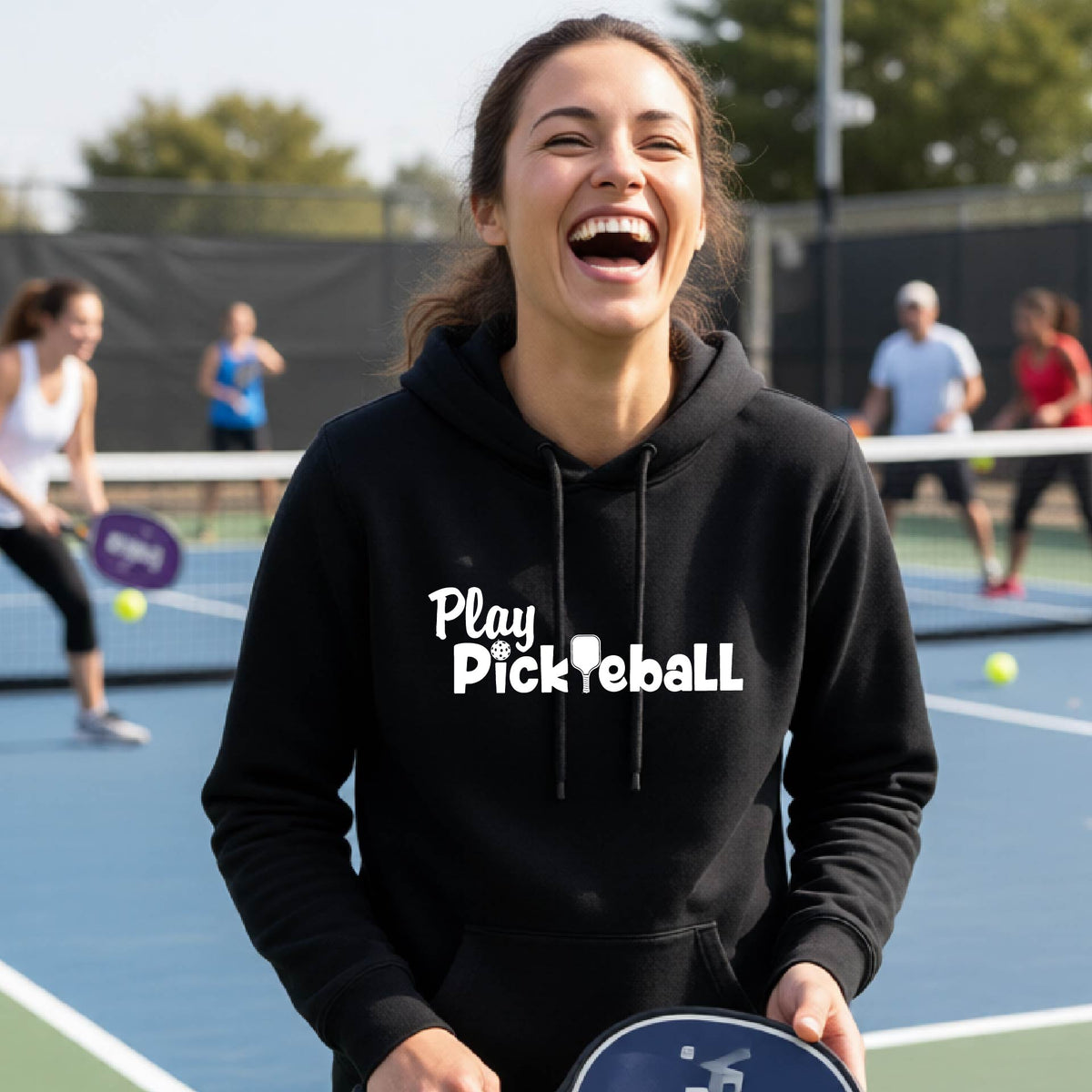 A woman laughing on a pickleball court wearing a black hoodie with the text 'Play Pickleball' in white. Other players and a blue pickleball court are visible in the background.