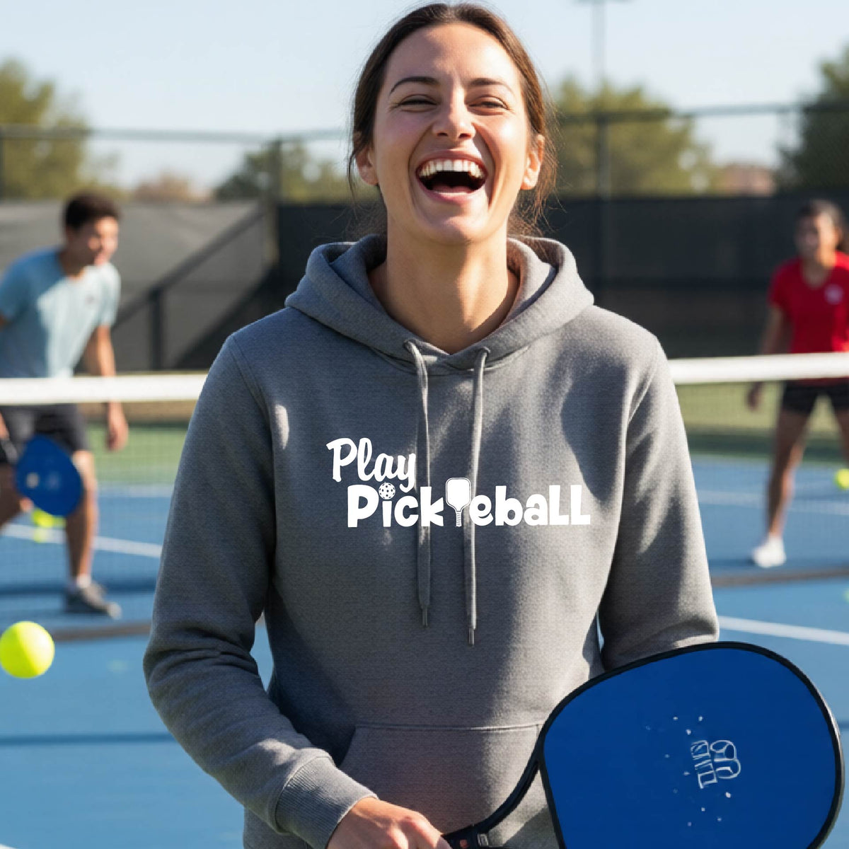 A woman laughing on a pickleball court wearing a heather gray hoodie with 'Play Pickleball' printed in white across the chest, holding a blue pickleball paddle.