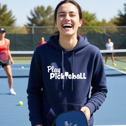 A woman laughing on an outdoor pickleball court wearing a navy blue hoodie with the white text 'Play Pickleball' across the chest, holding a pickleball paddle.