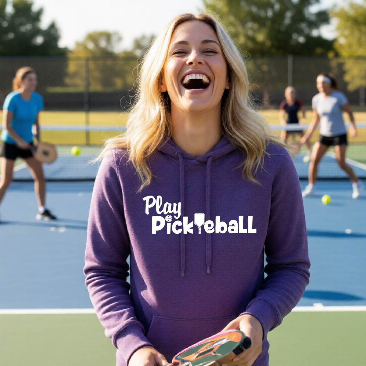 A woman laughing on an outdoor pickleball court wearing a purple hoodie with the text 'Play Pickleball' printed in white across the chest.