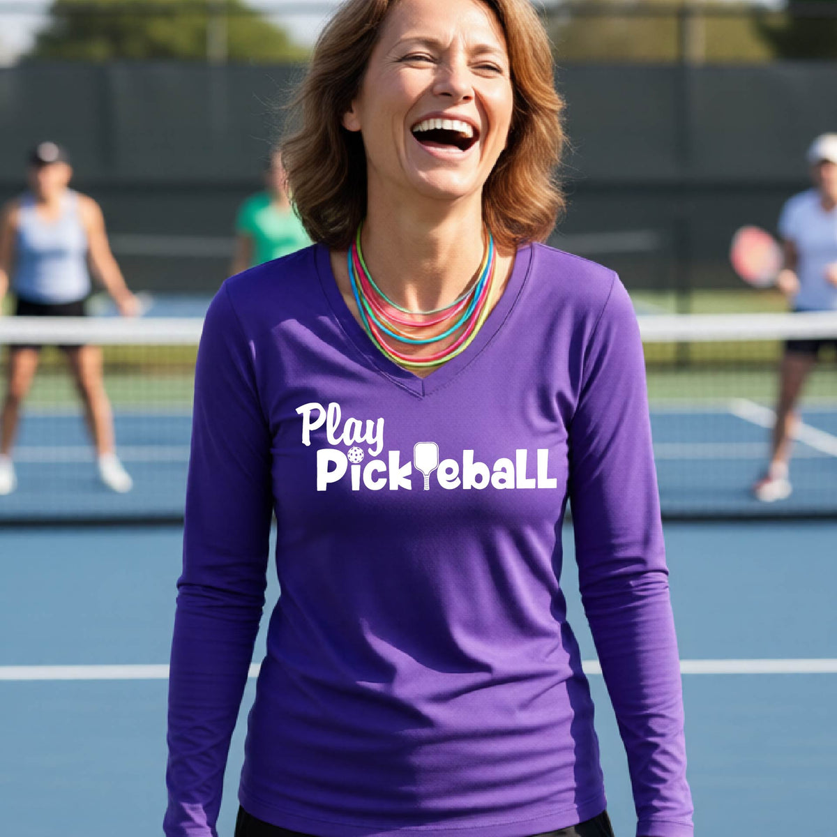 A woman laughing on a pickleball court wearing a purple long-sleeve shirt with 'Play Pickleball' printed in white across the chest.