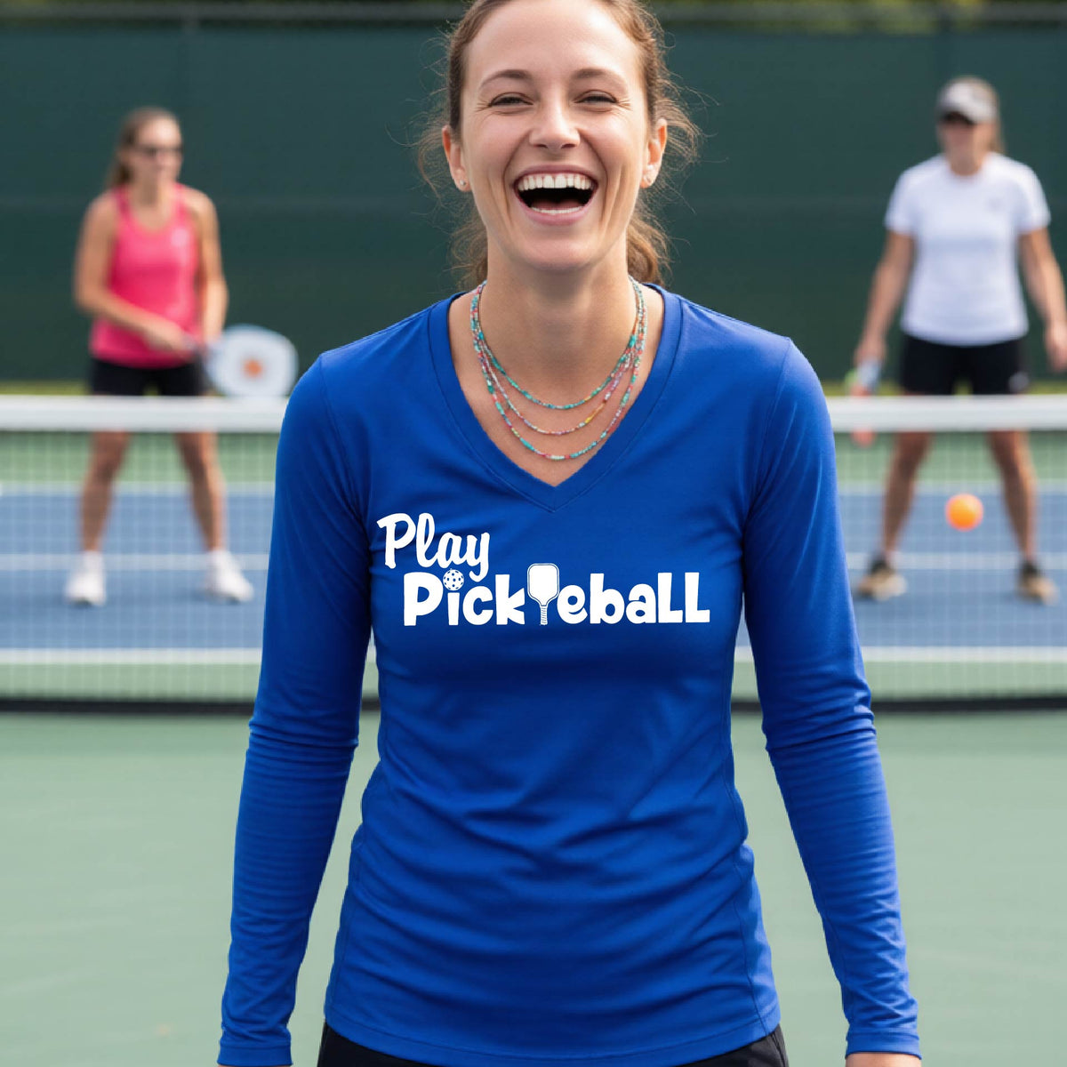 A woman laughing on a tennis court wearing a bright blue long-sleeve V-neck shirt with the white text 'Play Pickleball' across the chest. Two other players are blurred in the background near the net.