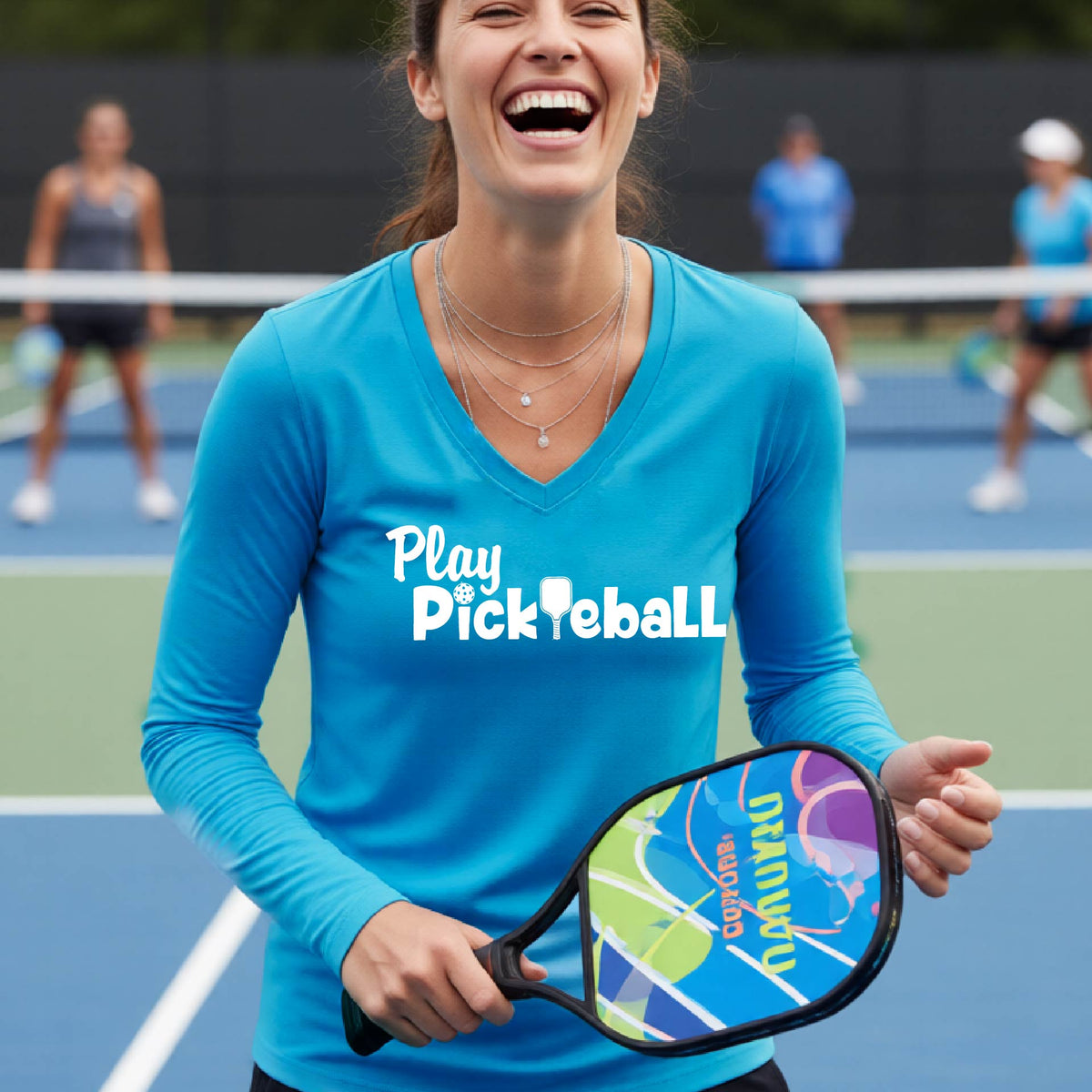 A woman on a pickleball court wearing a bright blue long-sleeve V-neck shirt with the text 'Play Pickleball' in white, holding a pickleball paddle. Other players are blurred in the background on the court.