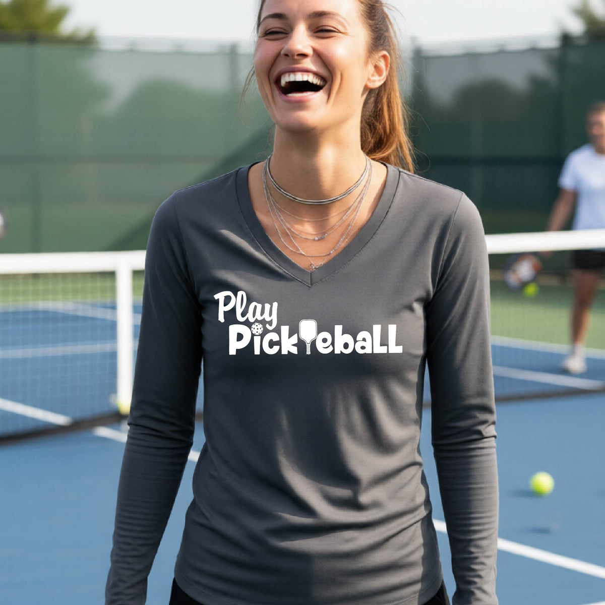 A woman laughing on a tennis court wearing a dark gray long-sleeve V-neck shirt with the white text 'Play Pickleball' printed across the chest.