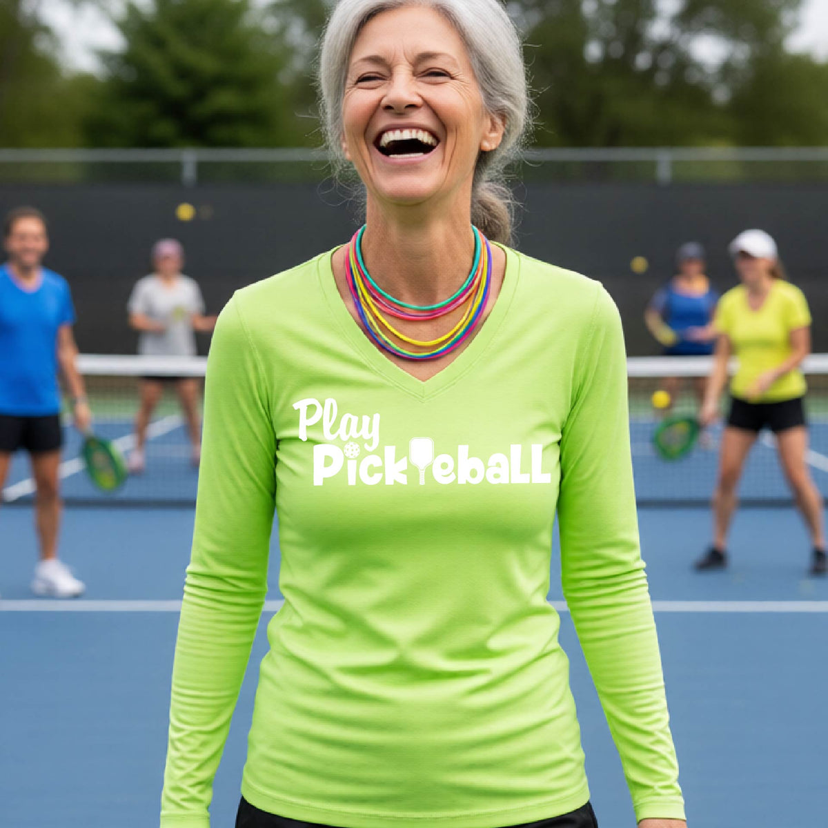 A smiling older woman stands on a blue pickleball court wearing a bright lime green long-sleeve shirt with the words 'Play Pickleball' printed in white across the chest. Other players are blurred in the background.