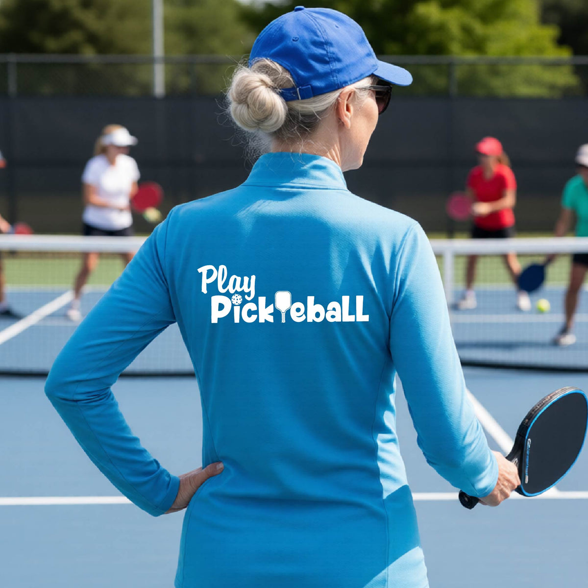 A woman on a pickleball court seen from behind, wearing a bright blue long-sleeve athletic shirt with the white text 'Play Pickleball' across the back, including a pickleball paddle icon in place of the L in pickleball. 