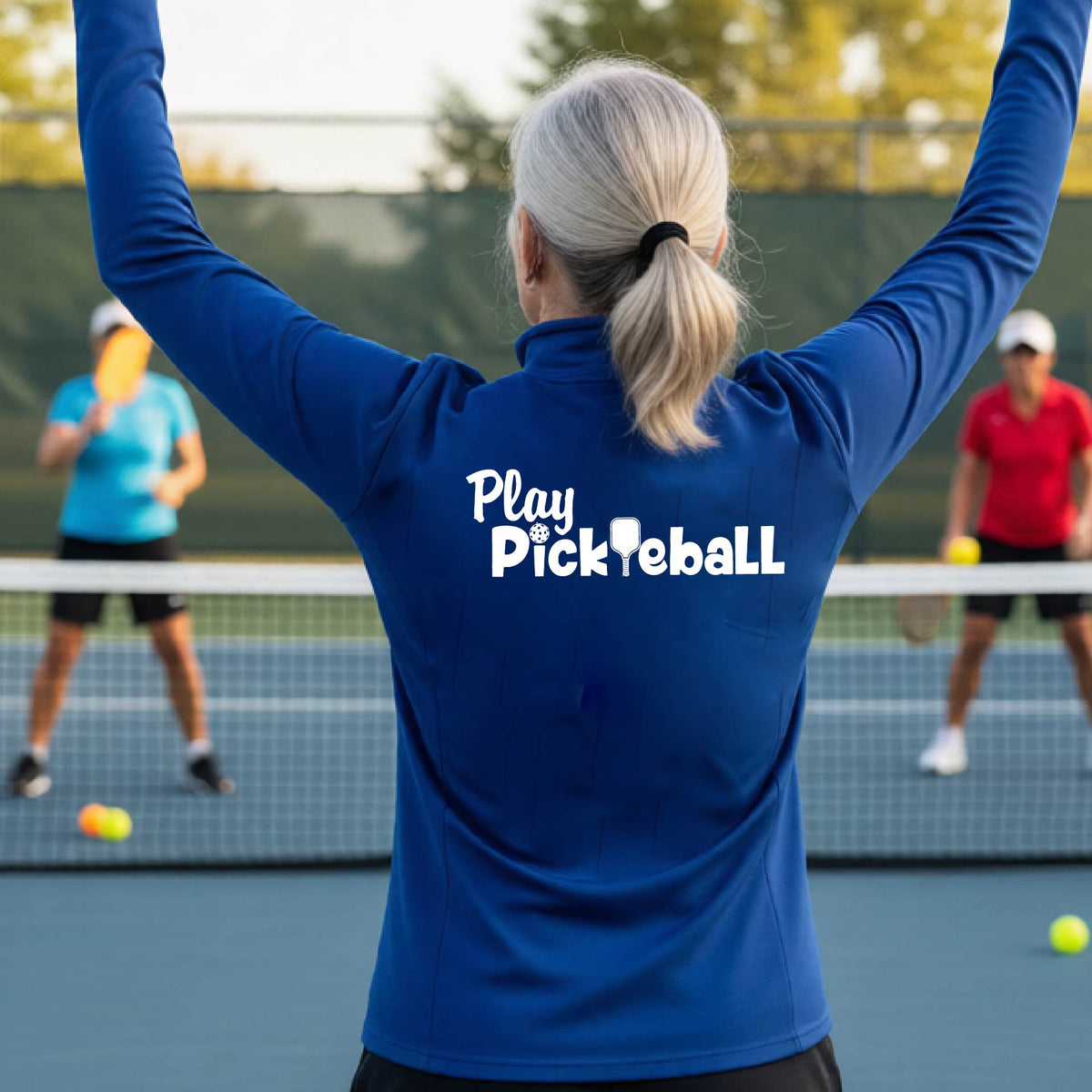 A woman on a pickleball court seen from behind, wearing a royal blue long-sleeve athletic shirt with the white text 'Play Pickleball' across the back, including a pickleball paddle icon in place of the L in pickleball. 
