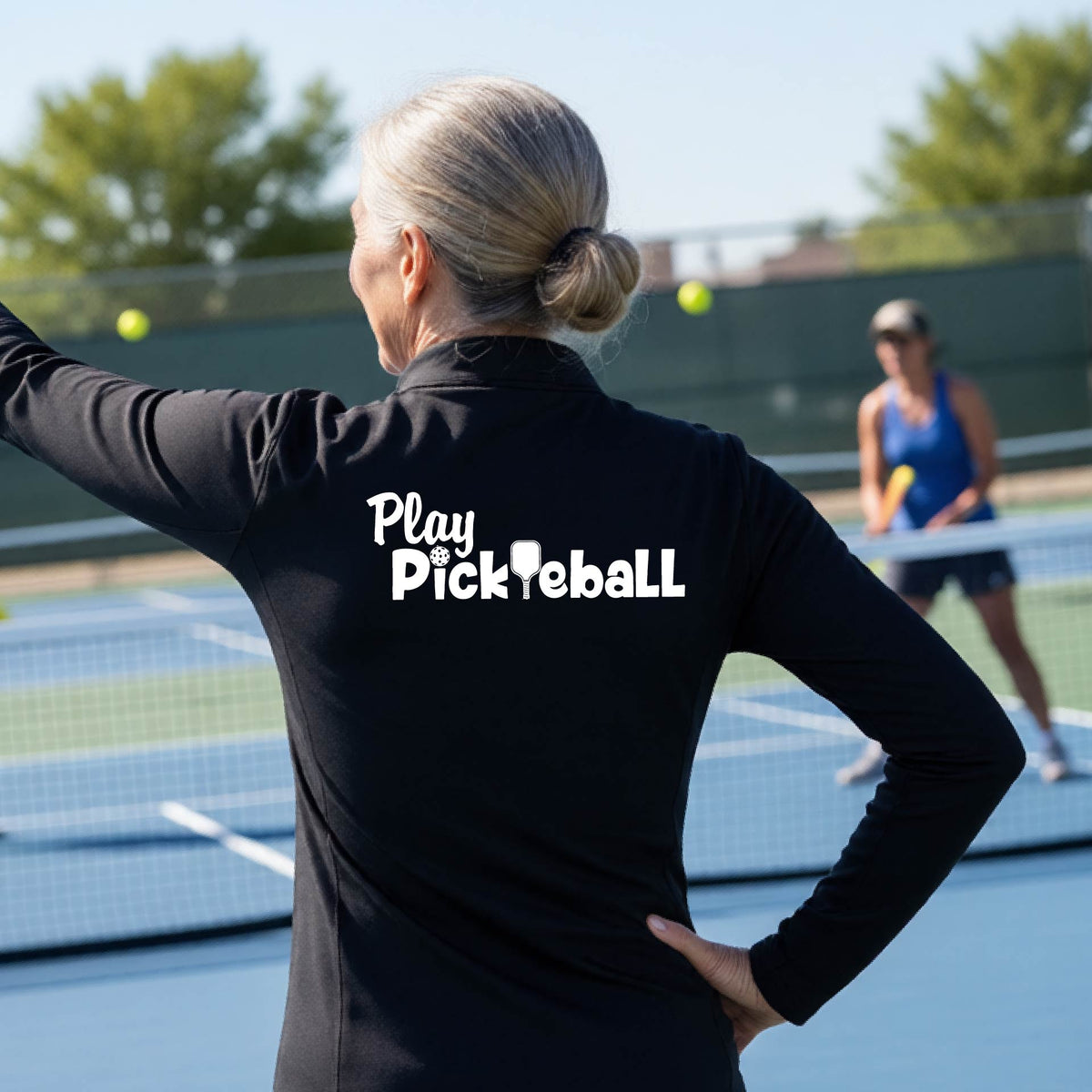 A woman on a pickleball court seen from behind, wearing a black long-sleeve athletic shirt with the white text 'Play Pickleball' across the back, including a pickleball paddle icon in place of the L in pickleball. 