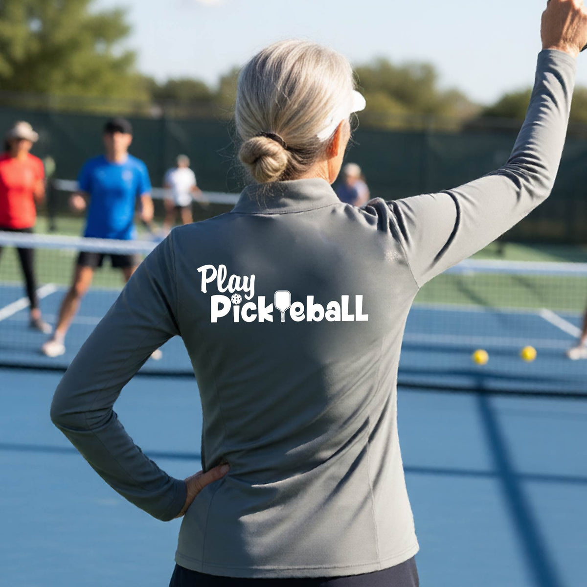A woman on a pickleball court seen from behind, wearing a gray long-sleeve athletic shirt with the white text 'Play Pickleball' across the back, including a pickleball paddle icon in place of the L in pickleball. 