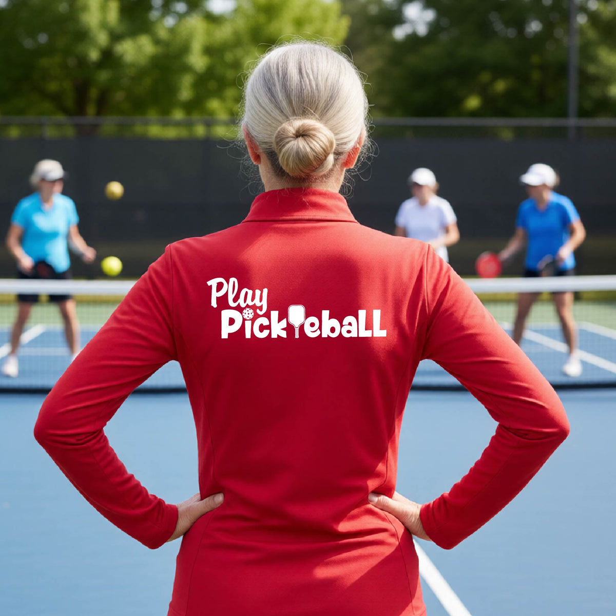 A woman on a pickleball court seen from behind, wearing a red long-sleeve athletic shirt with the white text 'Play Pickleball' across the back, including a pickleball paddle icon in place of the L in pickleball. 