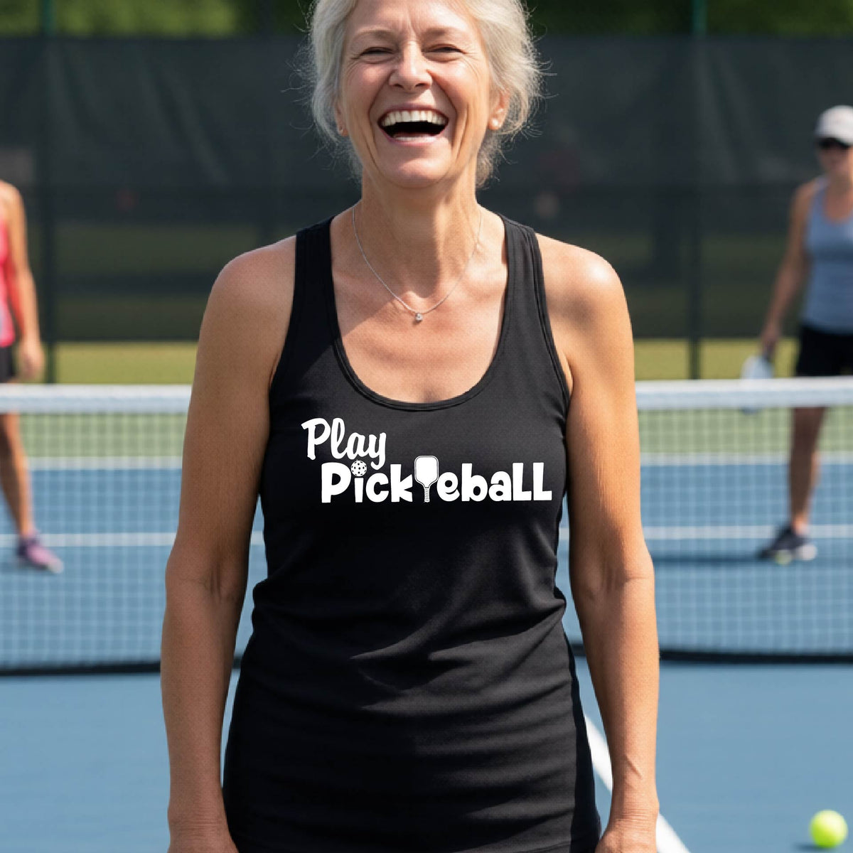 A woman on a pickleball court, wearing a black racerback tank top with the white text 'Play Pickleball' across the back, including a pickleball paddle icon in place of the L in pickleball. 
