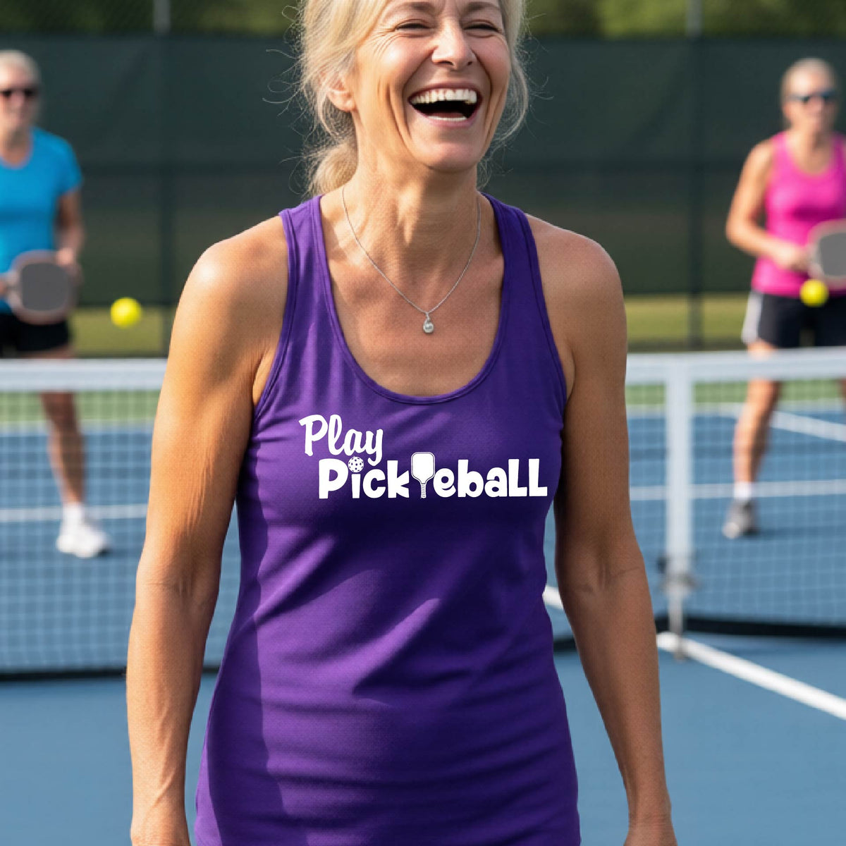 A woman on a pickleball court, wearing a purple racerback tank top with the white text 'Play Pickleball' across the back, including a pickleball paddle icon in place of the L in pickleball. 
