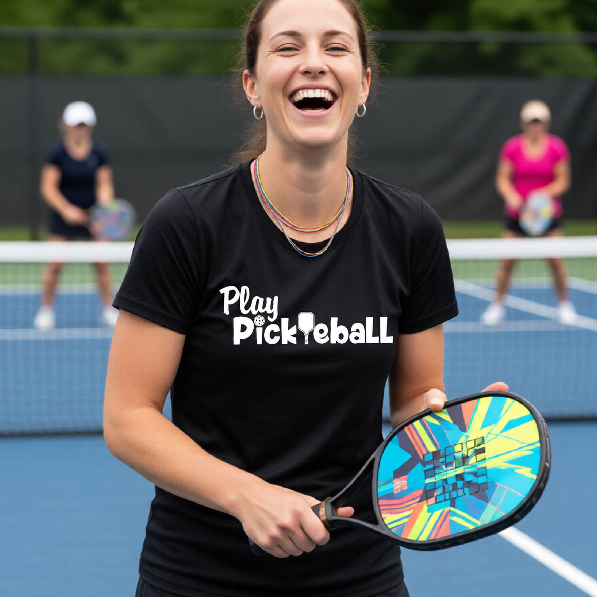 A woman laughing on a pickleball court wearing a black t-shirt with 'Play Pickleball' printed in white, holding a colorful paddle. Two other players are blurred in the background on the court.