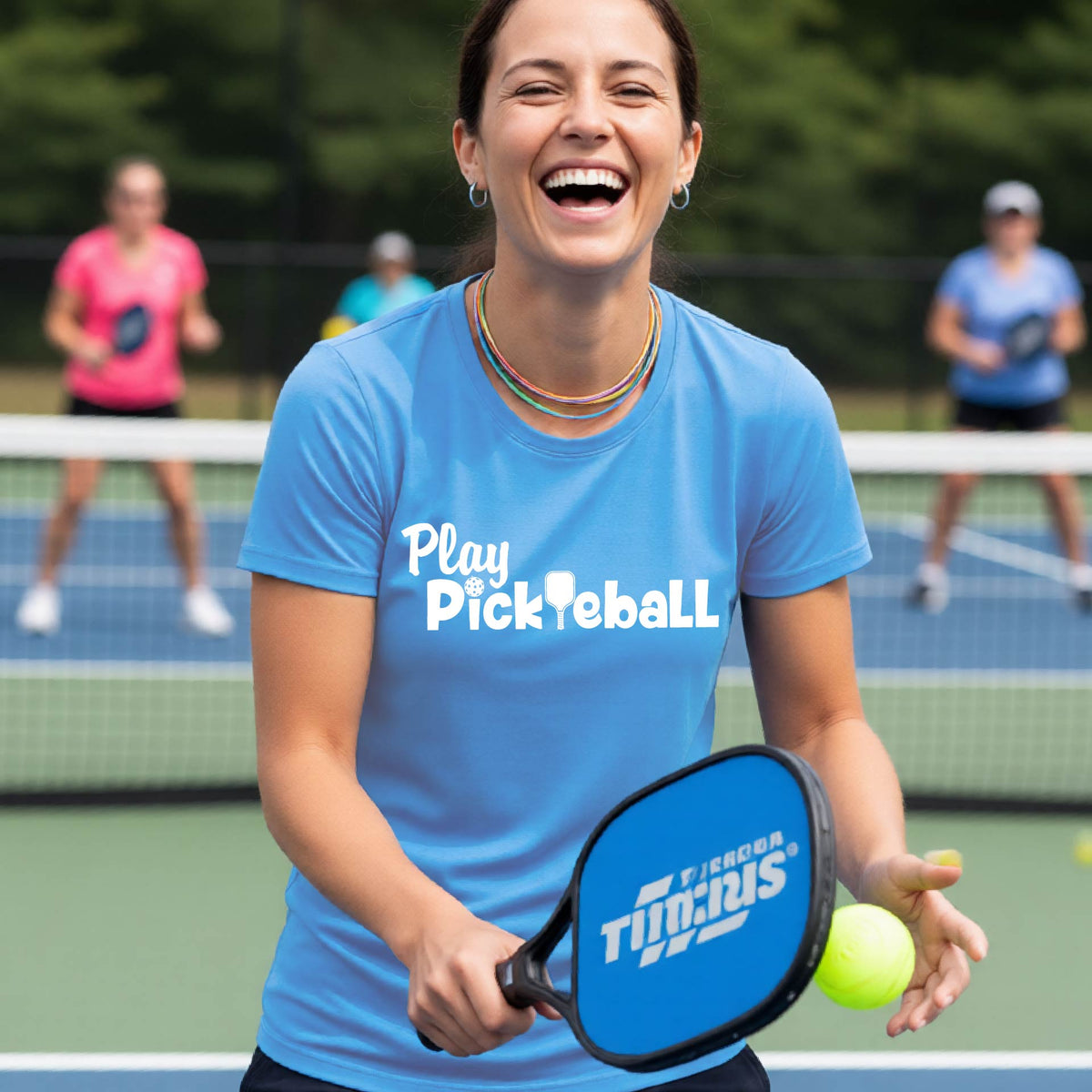 A woman on a pickleball court wearing a bright blue T-shirt with the text 'Play Pickleball' in white, holding a blue and black Trixus paddle and a yellow tennis ball, with other players blurred in the background.