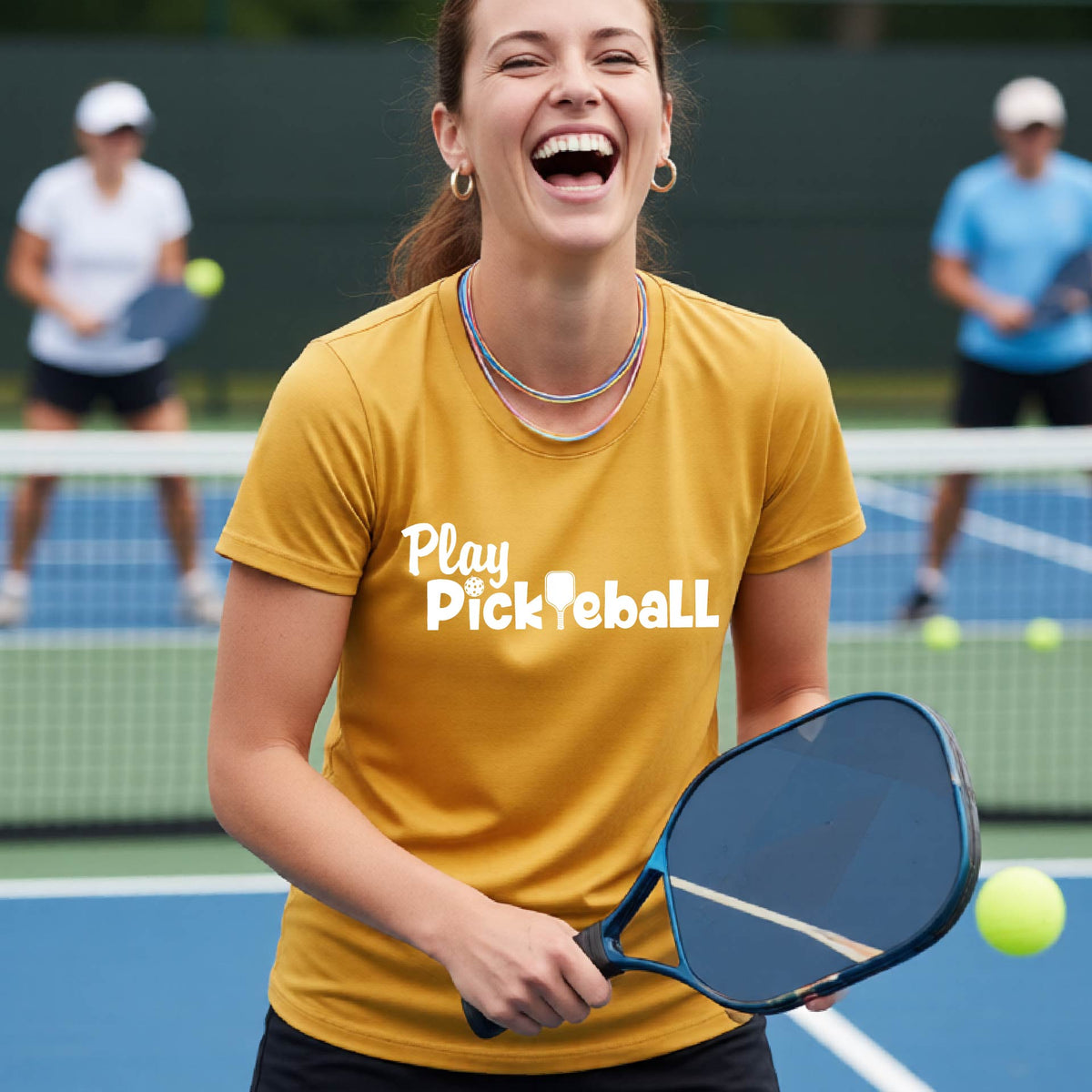 A woman laughing on a pickleball court wearing a mustard yellow t-shirt with the white text 'Play Pickleball' across the chest, holding a paddle and a yellow ball in the foreground, with other players blurred in the background.
