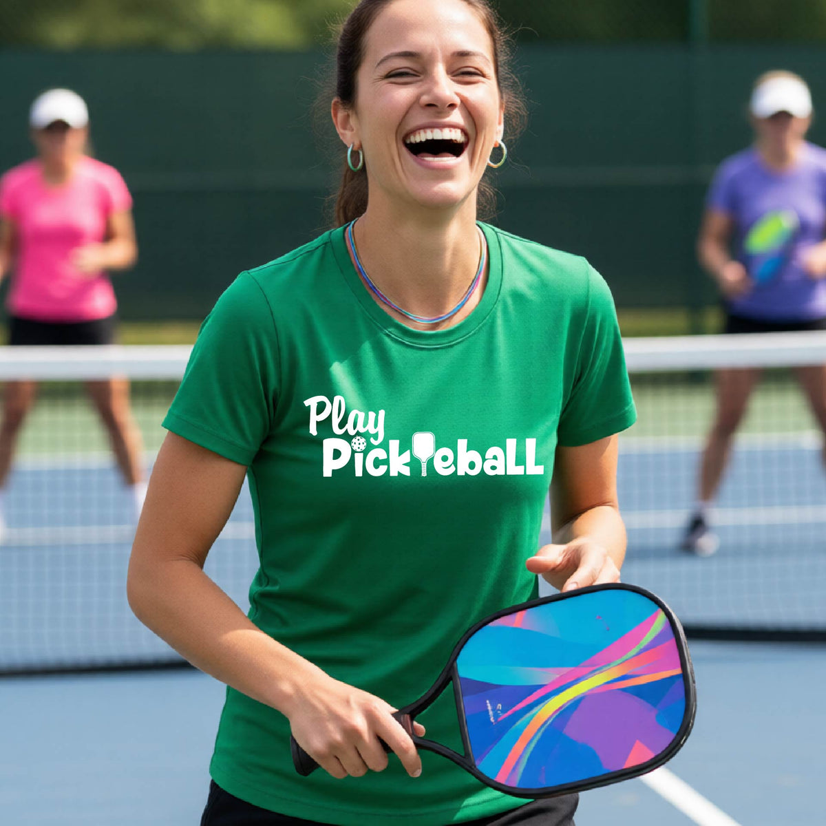 A woman laughing on a pickleball court wearing a green t-shirt with the text 'Play Pickleball' in white, holding a colorful paddle. Two other players are blurred in the background near the net.