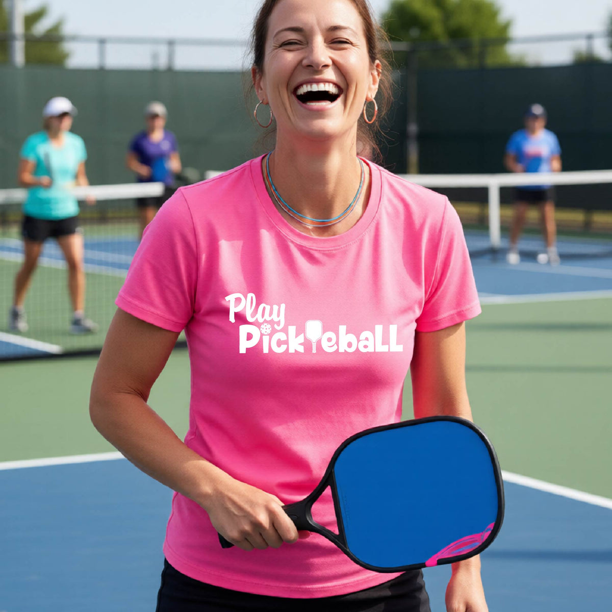 A woman laughing on a pickleball court wearing a bright pink t-shirt with the text 'Play Pickleball' in white,.