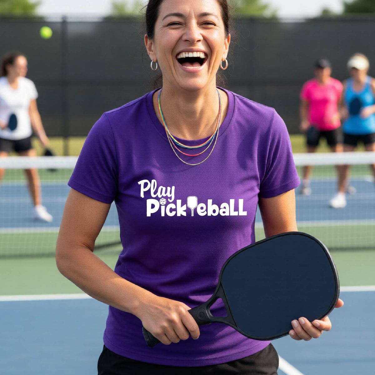 A woman laughing on an outdoor pickleball court wearing a purple t-shirt with 'Play Pickleball' printed in white, holding a pickleball paddle.