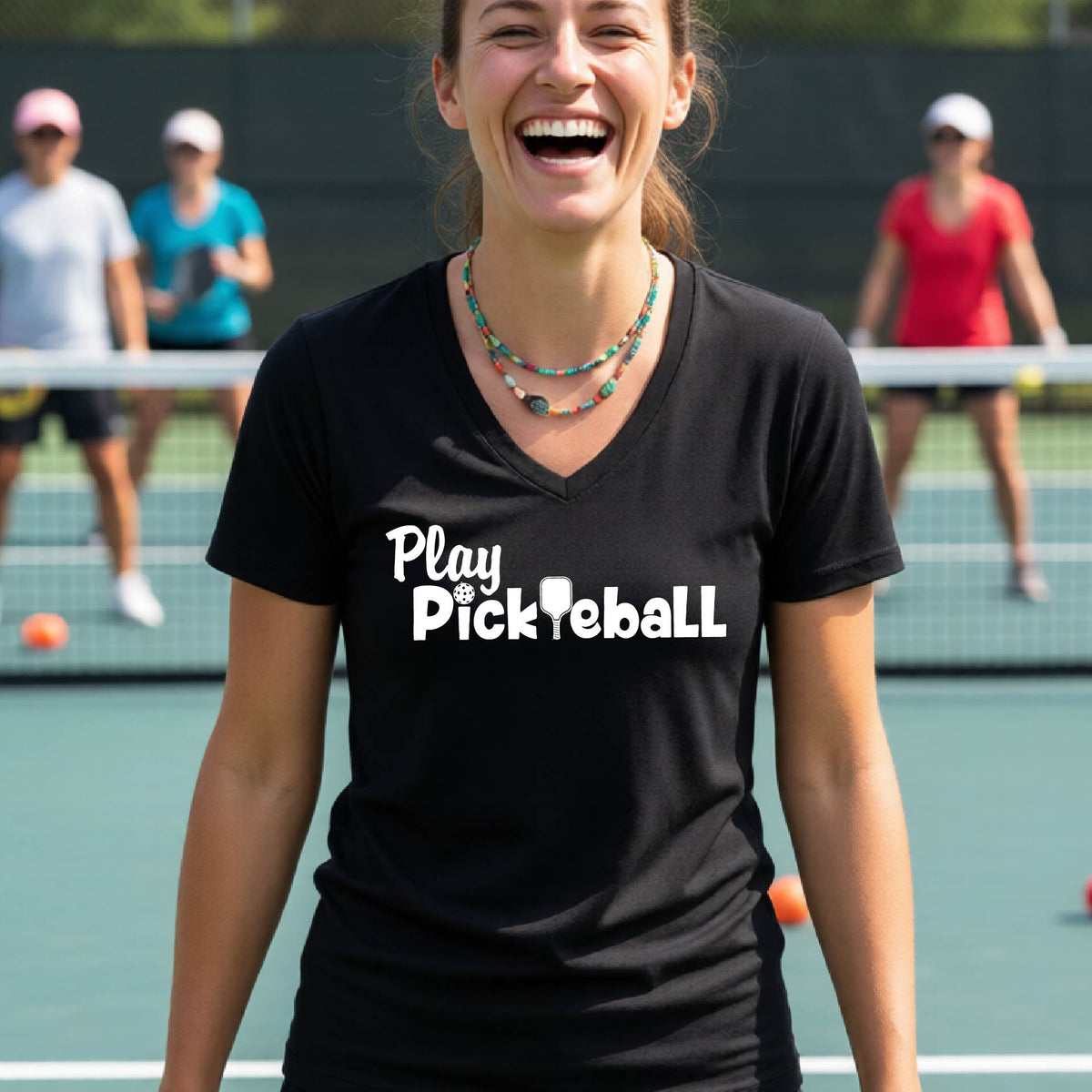 A woman laughing on a pickleball court wearing a black V-neck t-shirt with 'Play Pickleball' printed in white across the chest.
