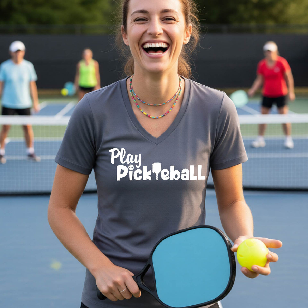 A woman laughing on a pickleball court wearing a gray V-neck t-shirt with 'Play Pickleball' printed in white across the chest.