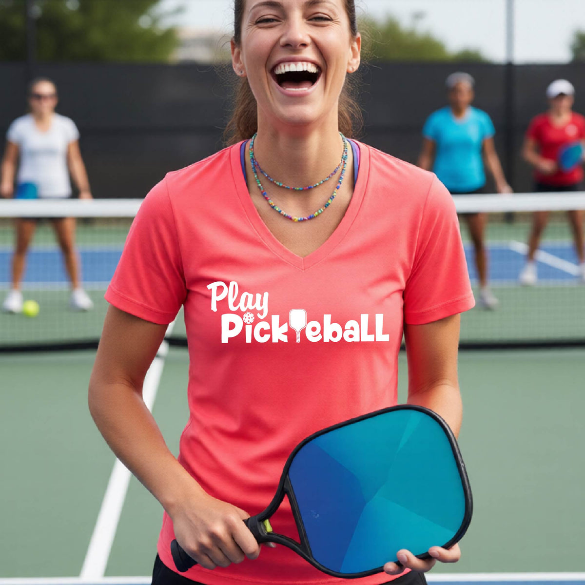A woman laughing on a pickleball court wearing a coral pink V-neck t-shirt with 'Play Pickleball' printed in white across the chest.