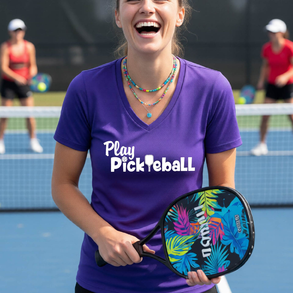 A woman laughing on a pickleball court wearing a purple V-neck t-shirt with 'Play Pickleball' printed in white across the chest.
