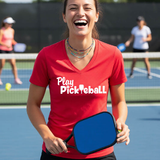 A woman laughing on a pickleball court wearing a red V-neck t-shirt with 'Play Pickleball' printed in white across the chest.