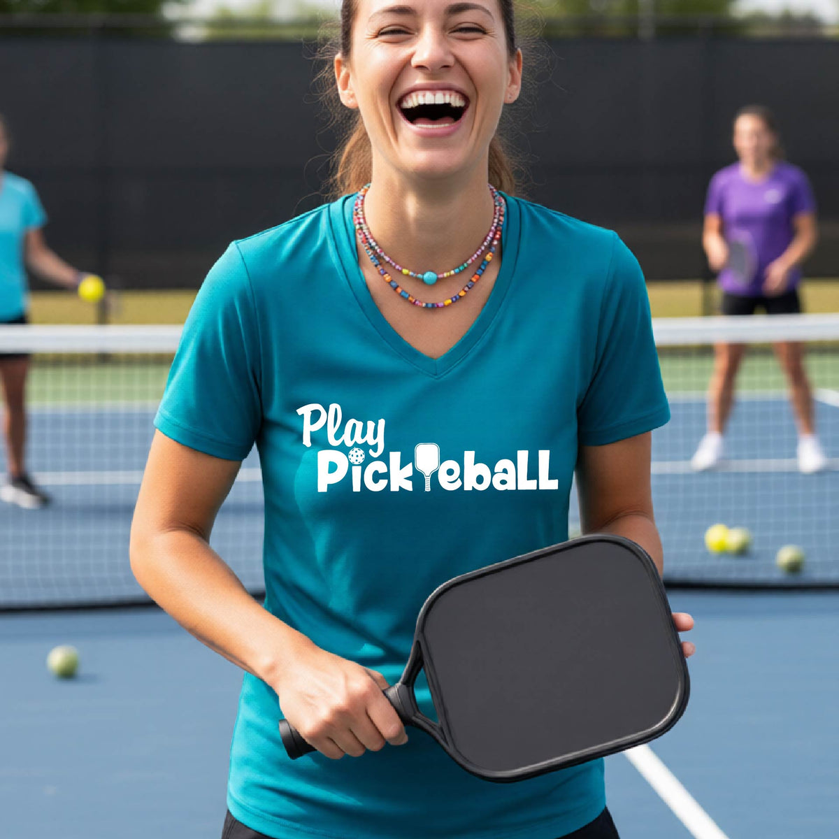 A woman laughing on a pickleball court wearing a teal V-neck t-shirt with 'Play Pickleball' printed in white across the chest.