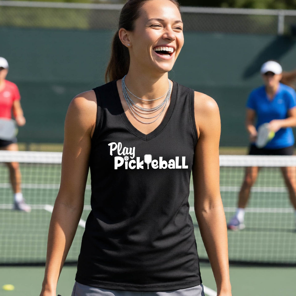 A woman laughing on a pickleball court wearing a black tank top with the white text 'Play Pickleball' across the chest. The background shows a green tennis court with other players blurred.