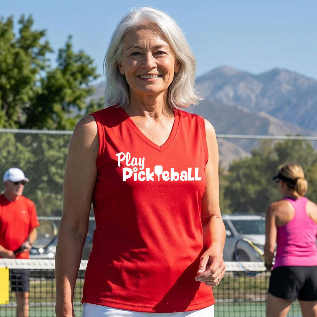 A smiling senior woman with white hair stands on a pickleball court wearing a bright red sleeveless V-neck shirt with the text 'Play Pickleball' in white. Other players and mountains are visible in the background.