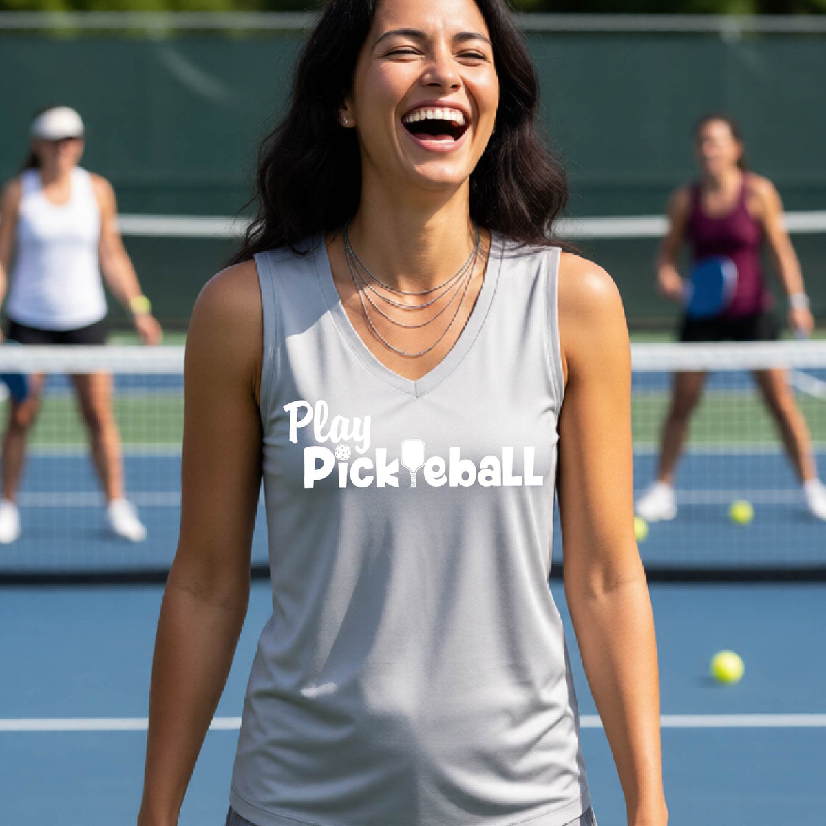 A woman laughing on a pickleball court wearing a light gray tank top with the text 'Play Pickleball' printed on the front. Two other players are blurred in the background behind the net.