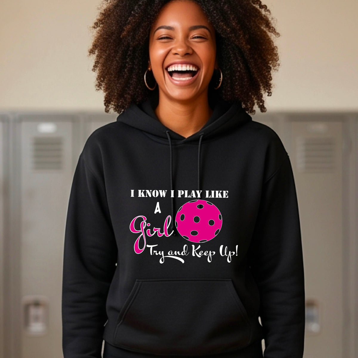 A woman laughing while wearing a black hoodie with a pink and white graphic that reads 'I KNOW I PLAY LIKE A Girl Try and Keep Up!' with a pink pickleball illustration. 