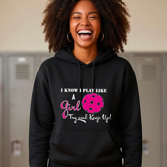 A woman laughing while wearing a black hoodie with a pink and white graphic that reads 'I KNOW I PLAY LIKE A Girl Try and Keep Up!' with a pink pickleball illustration. 