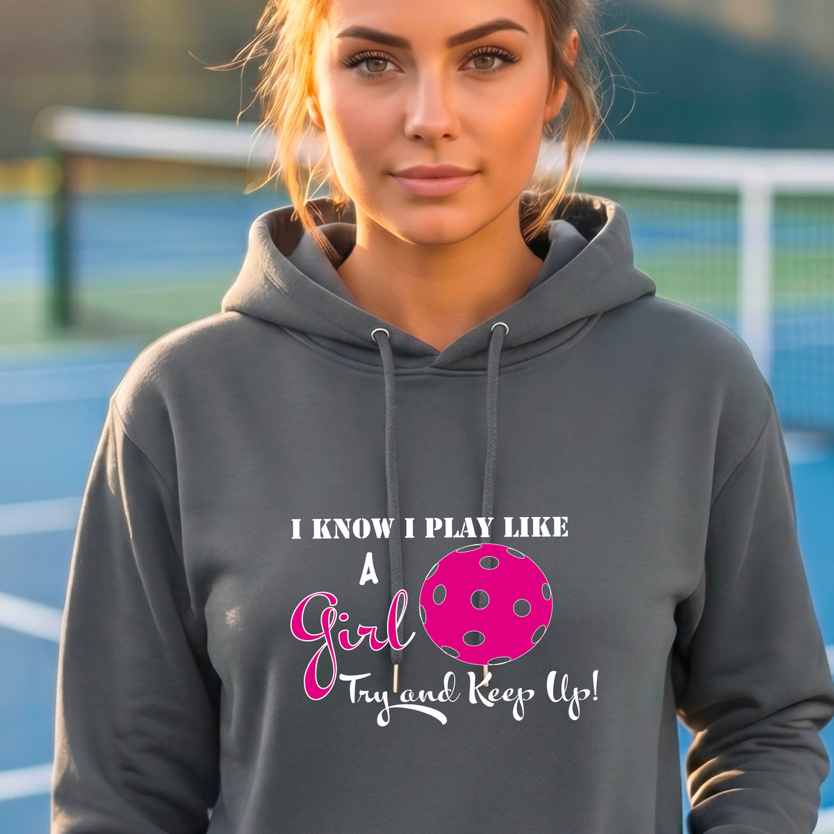 A woman smiling while wearing a gray hoodie with a pink and white graphic that reads 'I KNOW I PLAY LIKE A Girl Try and Keep Up!' with a pink pickleball illustration.
