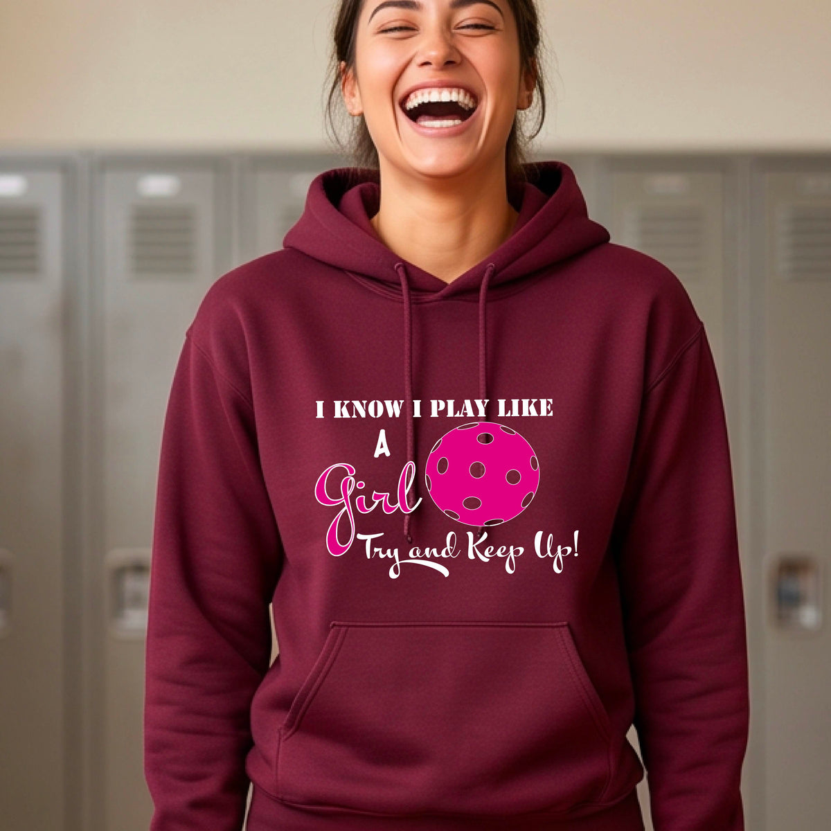 A woman laughing while wearing a maroon hoodie with a pink and white graphic that reads 'I KNOW I PLAY LIKE A Girl Try and Keep Up!' with a pink pickleball illustration.