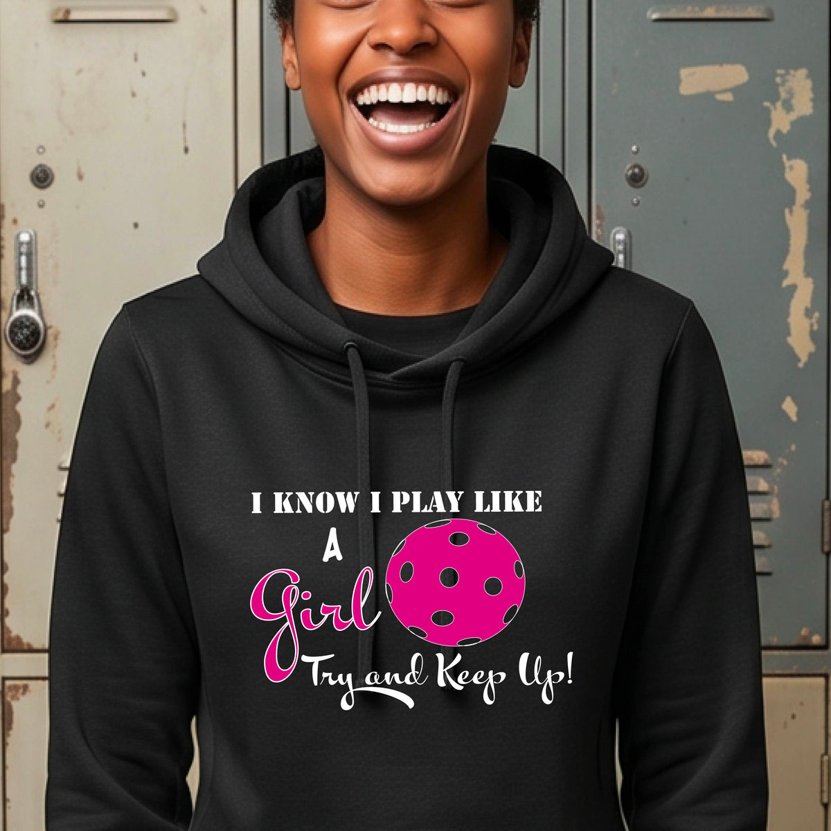 A close-up of a woman wearing a black cowl-neck hoodie pullover with a white graphic of ‘I KNOW I PLAY LIKE A GIRL TRY AND KEEP UP’ in white and pink with a pink pickleball icon, in front of school lockers. 