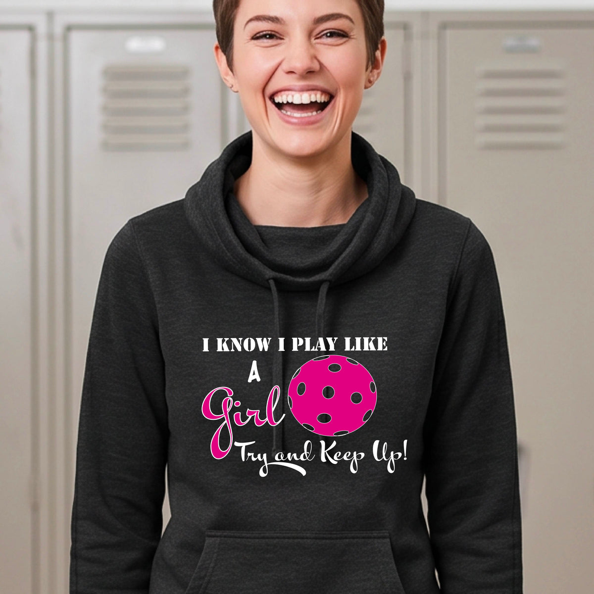 A close-up of a woman wearing a black cowl-neck hoodie pullover with a white graphic of ‘I KNOW I PLAY LIKE A GIRL TRY AND KEEP UP’ in white and pink with a pink pickleball icon, in front of a pickleball court. 