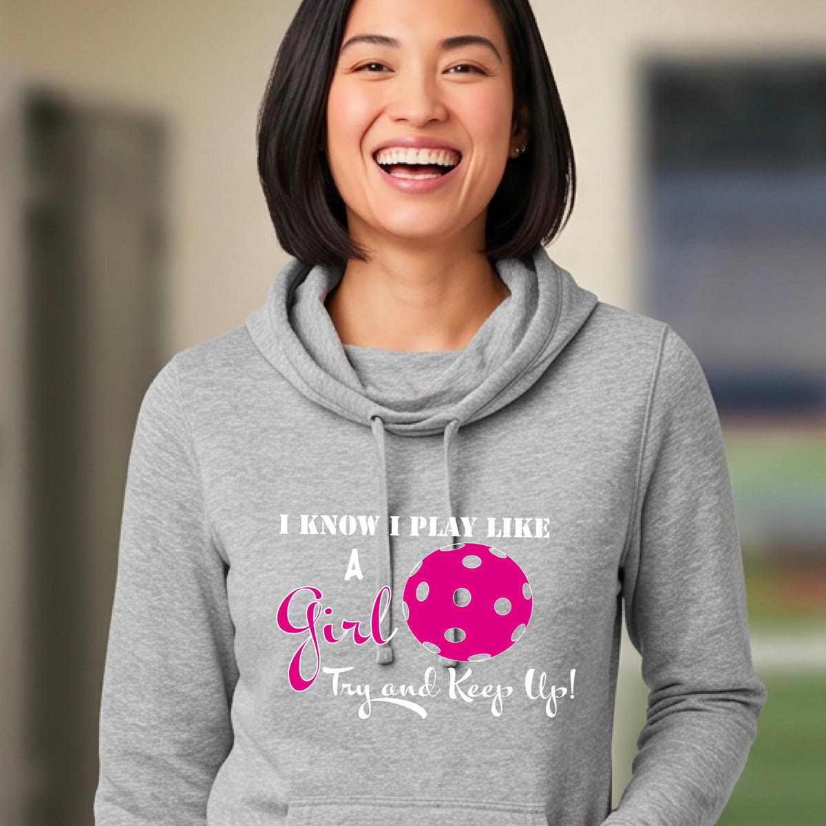 A close-up of a woman wearing a gray cowl-neck hoodie pullover with a white graphic of ‘I KNOW I PLAY LIKE A GIRL TRY AND KEEP UP’ in white and pink with a pink pickleball icon, in front of a pickleball court. 
