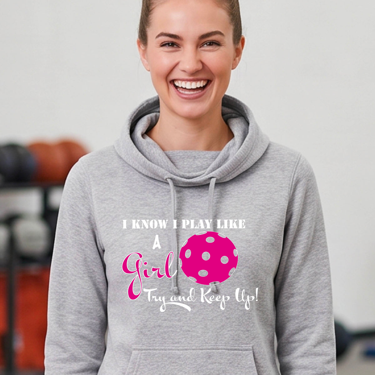 A close-up of a woman wearing a gray cowl-neck hoodie pullover with a white graphic of ‘I KNOW I PLAY LIKE A GIRL TRY AND KEEP UP’ in white and pink with a pink pickleball icon, in front of a pickleball court. 