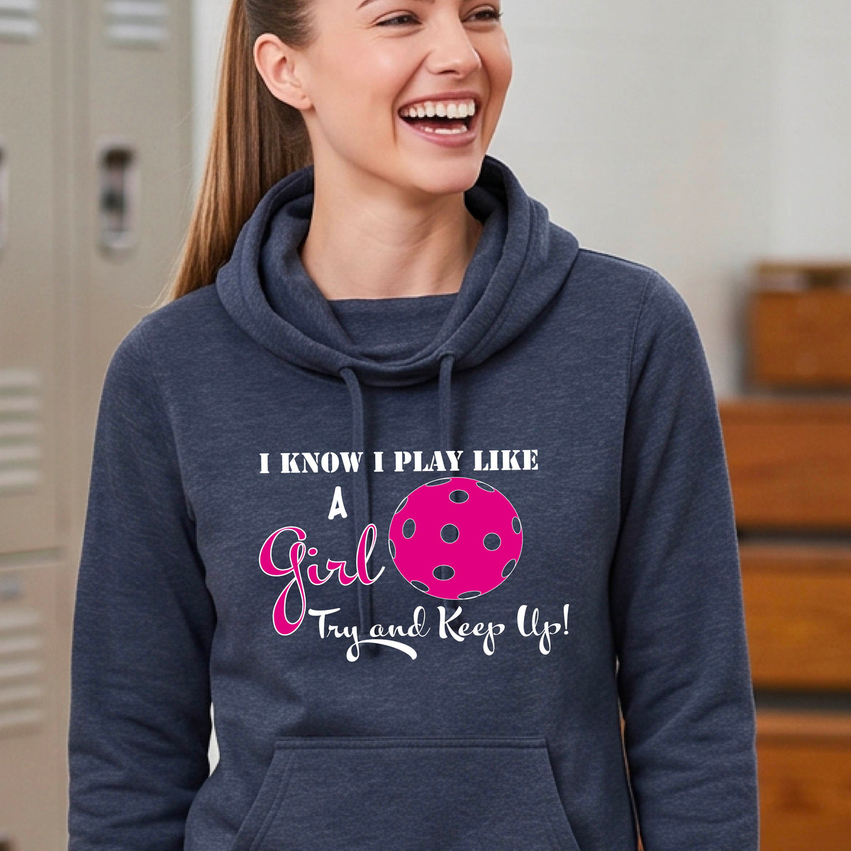A close-up of a woman wearing a navy blue cowl-neck hoodie pullover with a white graphic of ‘I KNOW I PLAY LIKE A GIRL TRY AND KEEP UP’ in white and pink with a pink pickleball icon, in front of a pickleball court. 
