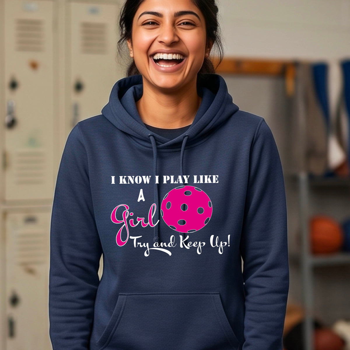 A close-up of a woman wearing a navy blue cowl-neck hoodie pullover with a white graphic of ‘I KNOW I PLAY LIKE A GIRL TRY AND KEEP UP’ in white and pink with a pink pickleball icon, in front of a pickleball court. 