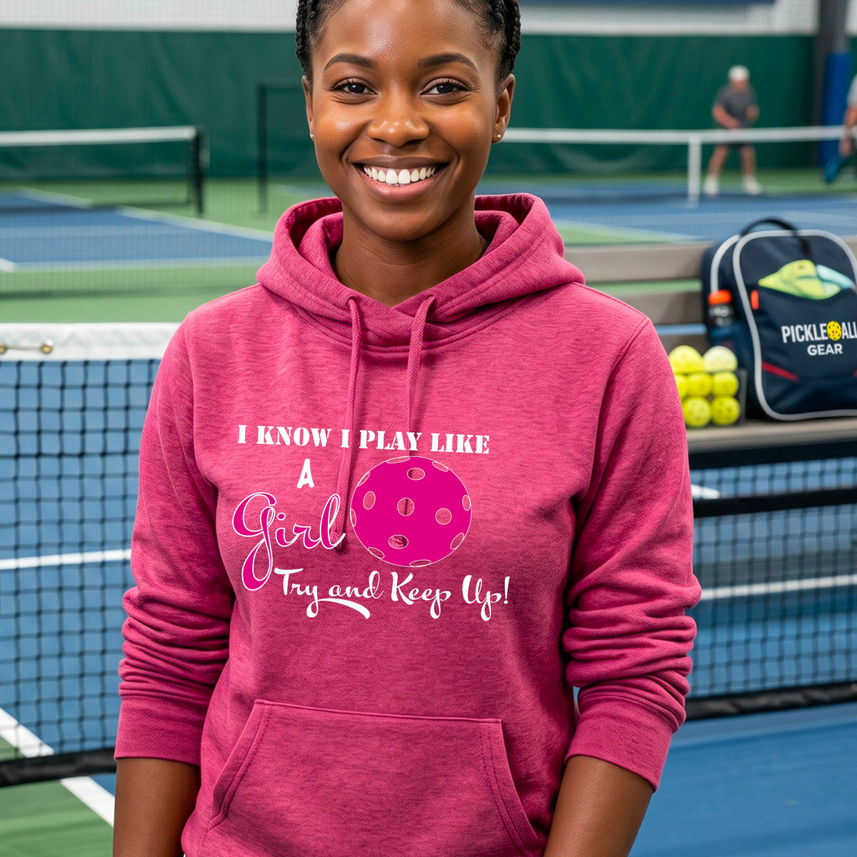 A close-up of a woman wearing a pink cowl-neck hoodie pullover with a white graphic of ‘I KNOW I PLAY LIKE A GIRL TRY AND KEEP UP’ in white and pink with a pink pickleball icon, in front of a pickleball court. 
