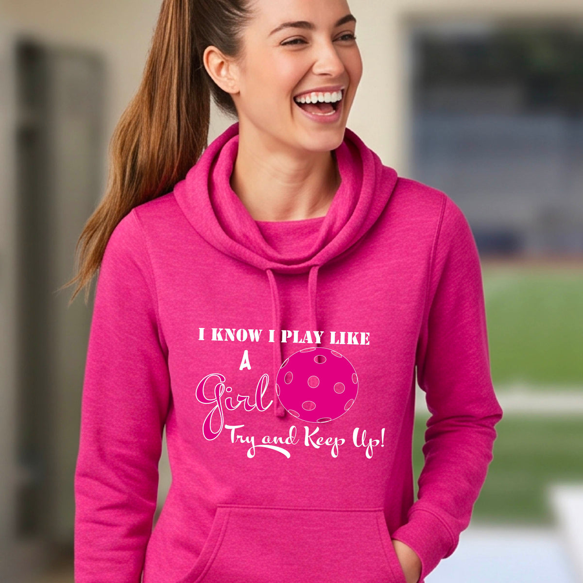 A close-up of a woman wearing a pink cowl-neck hoodie pullover with a white graphic of ‘I KNOW I PLAY LIKE A GIRL TRY AND KEEP UP’ in white and pink with a pink pickleball icon, in front of a pickleball court. 