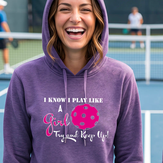 A close-up of a woman wearing a purple fitted hoodie pullover with a white graphic of ‘I KNOW I PLAY LIKE A GIRL TRY AND KEEP UP’ in white and pink with a pink pickleball icon, in front of a pickleball court. 