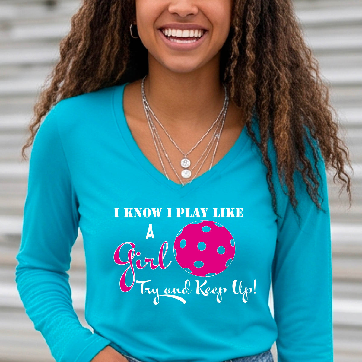 A woman laughing while wearing a bright blue  long-sleeve v-neck  t-shirt with a pink and white graphic that reads 'I KNOW I PLAY LIKE A Girl Try and Keep Up!' with a pink pickleball illustration.
